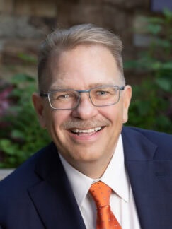 John Imig is a man with short light-colored hair and wearing glasses and an orange tie.