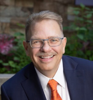 John Imig is a man with short light-colored hair and wearing glasses and an orange tie.