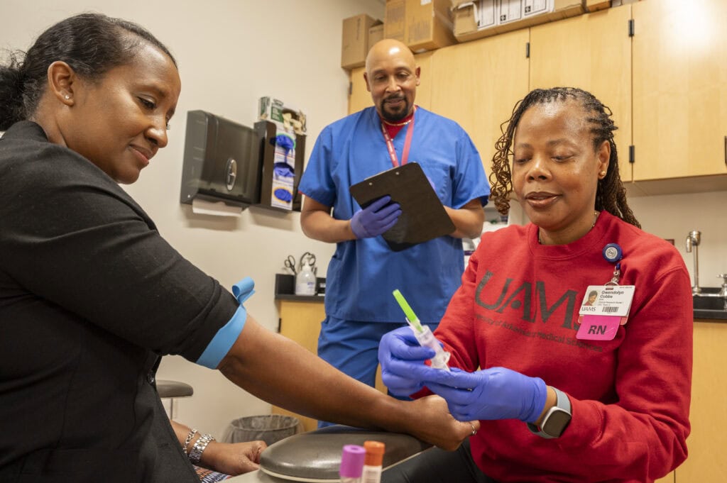 Photo shows three people in an exam room with one preparing a needle for injection and another holding a clipboard. 