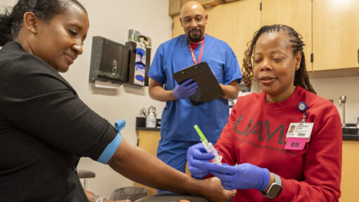 Photo shows three people in an exam room with one preparing a needle for injection and another holding a clipboard.