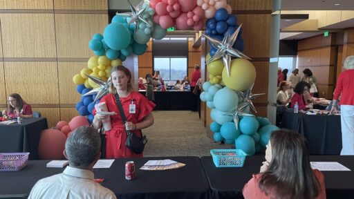 A woman talks to two people seated at a table. A arch of balloons over a doorway is in the background.