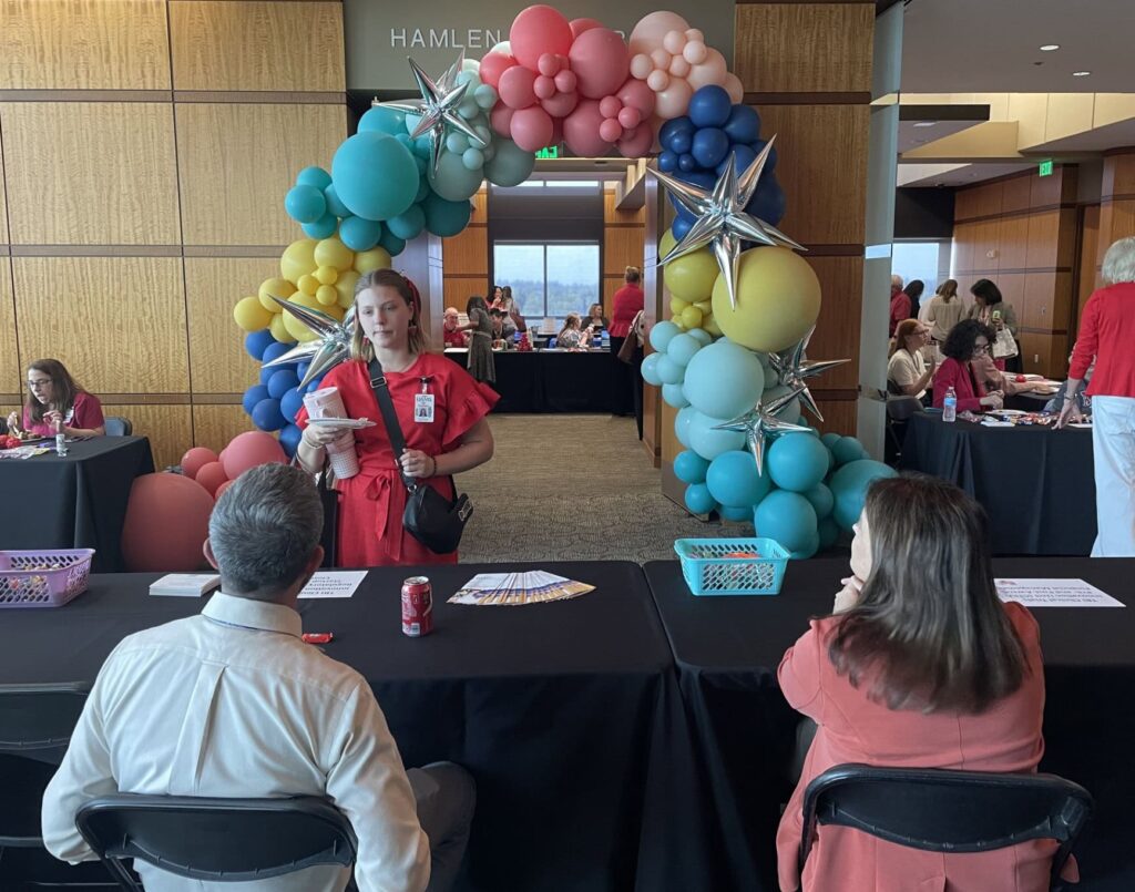 A woman talks to two people seated at a table. A arch of balloons over a doorway is in the background.